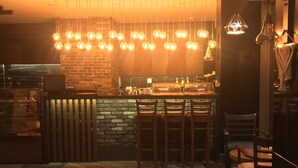 Zona de bar moderna con mostrador de madera, varias lámparas colgantes y un área de estar con sillas y una mesa.