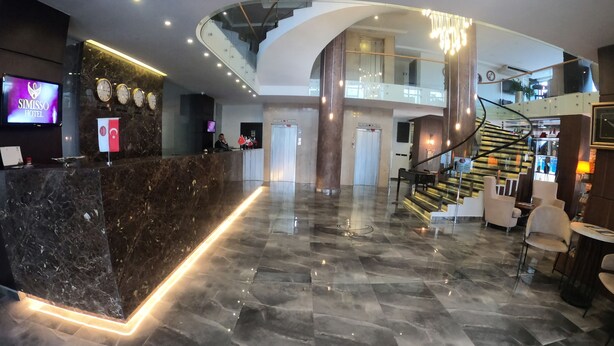 Un moderno vestíbulo de hotel con un mostrador de recepción de mármol, una pantalla digital, una escalera y un área de descanso.