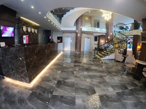 Een moderne hotellobby met een marmeren receptiebalie, een digitaal scherm, een trap en een zithoek.