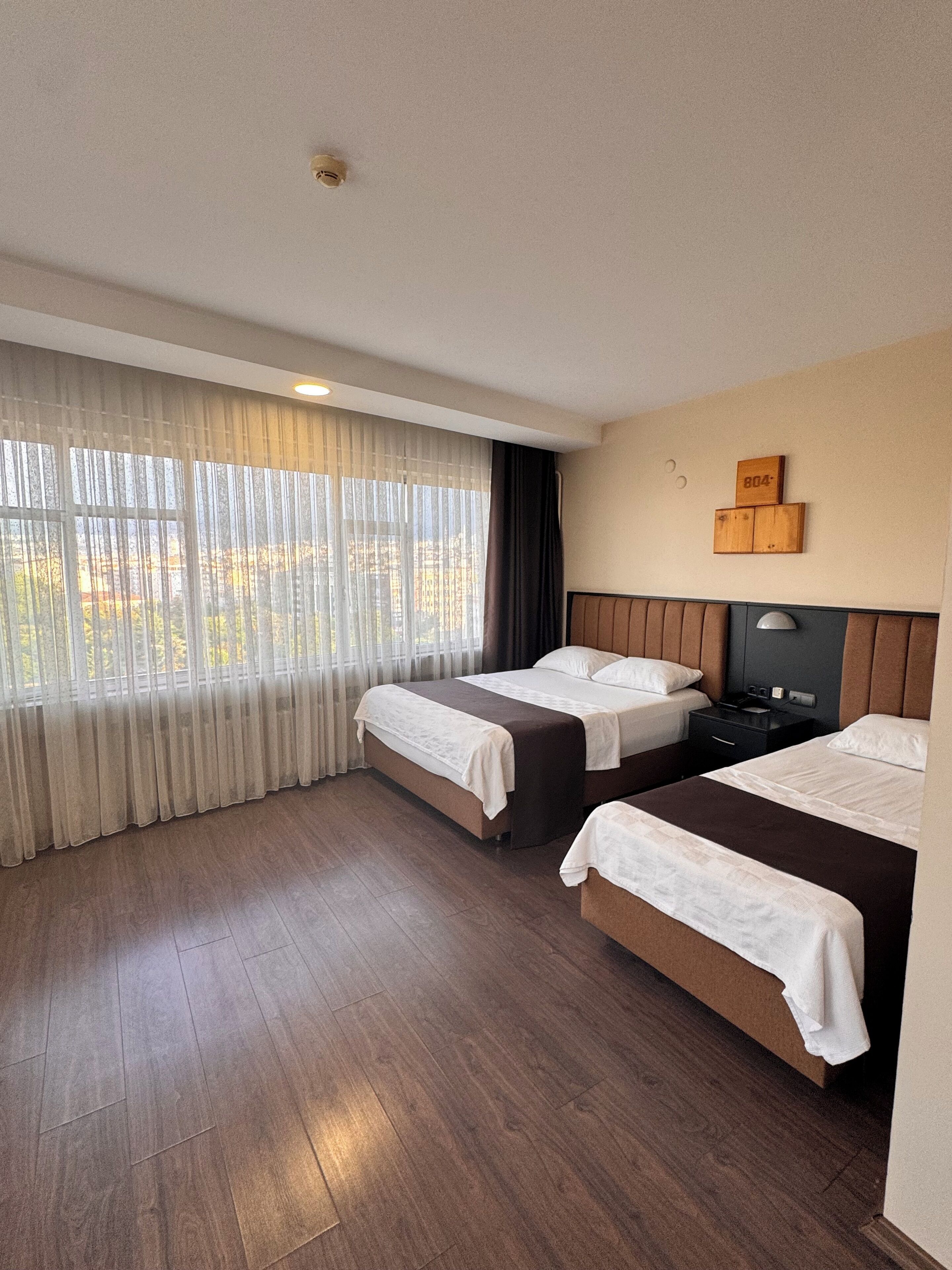 A hotel room with two beds, a wooden floor, a window with sheer curtains, and a wall-mounted light fixture.