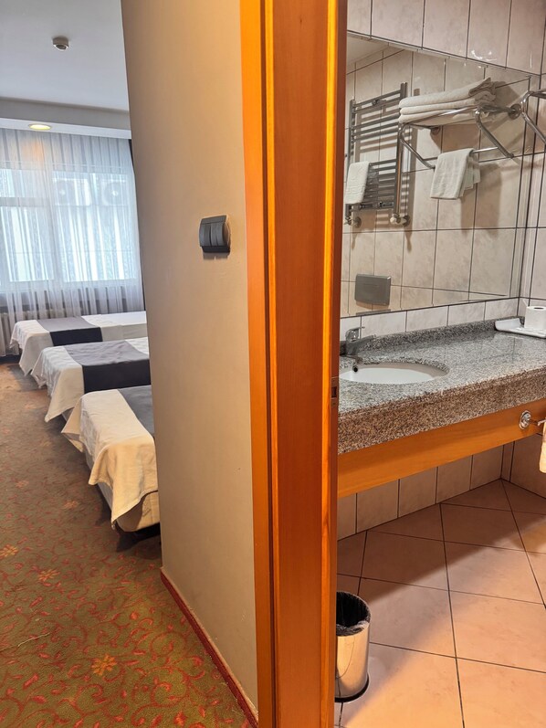 Habitación de hotel con baño que incluye un lavamanos con encimera de granito, un perchero y un espejo. Se observan camas con ropa blanca y una alfombra estampada.