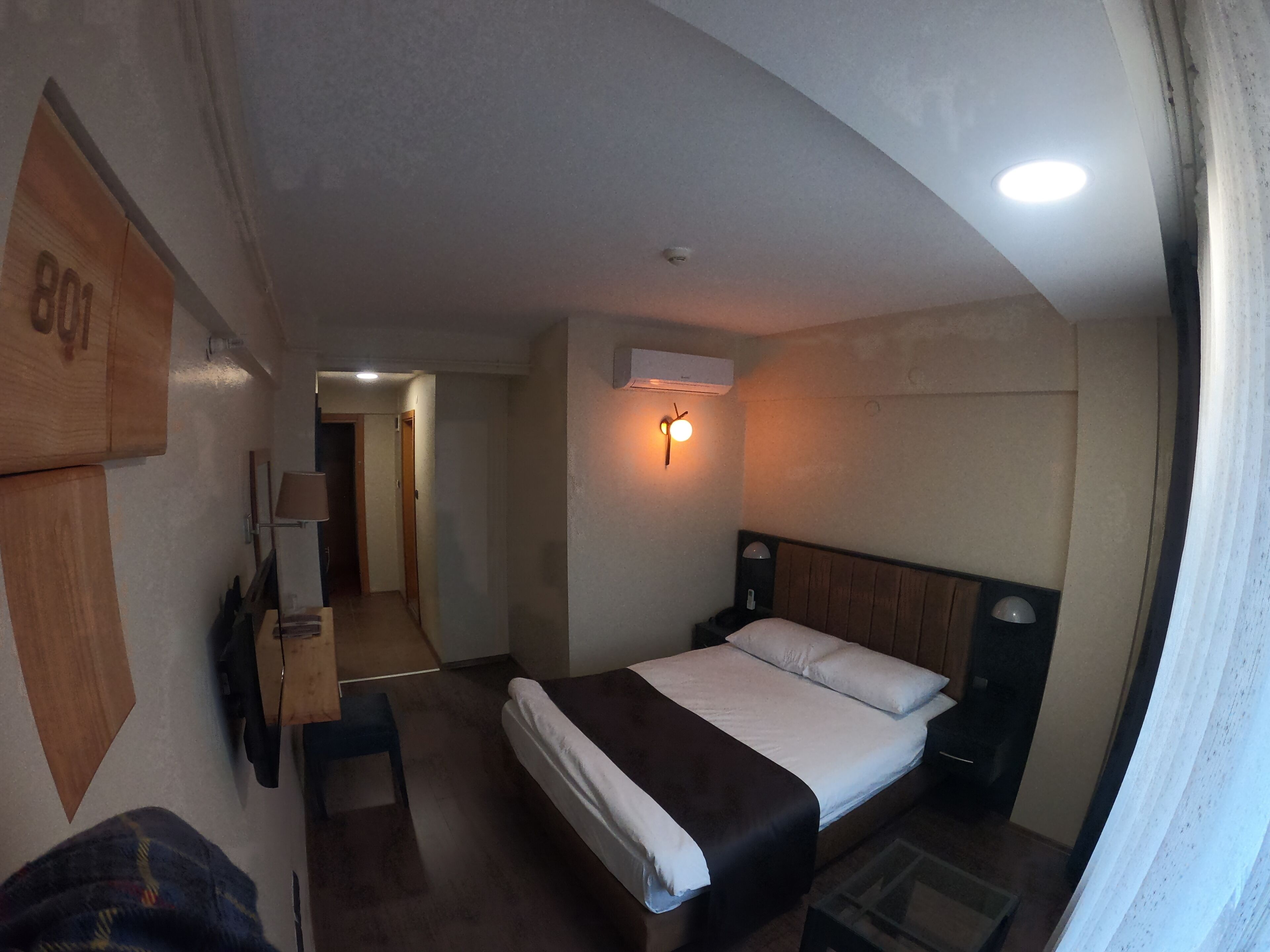 A hotel room with a bed, bedside table, lamp, and an air conditioning unit.