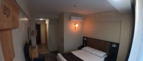 A hotel room with a bed, bedside table, lamp, and an air conditioning unit.