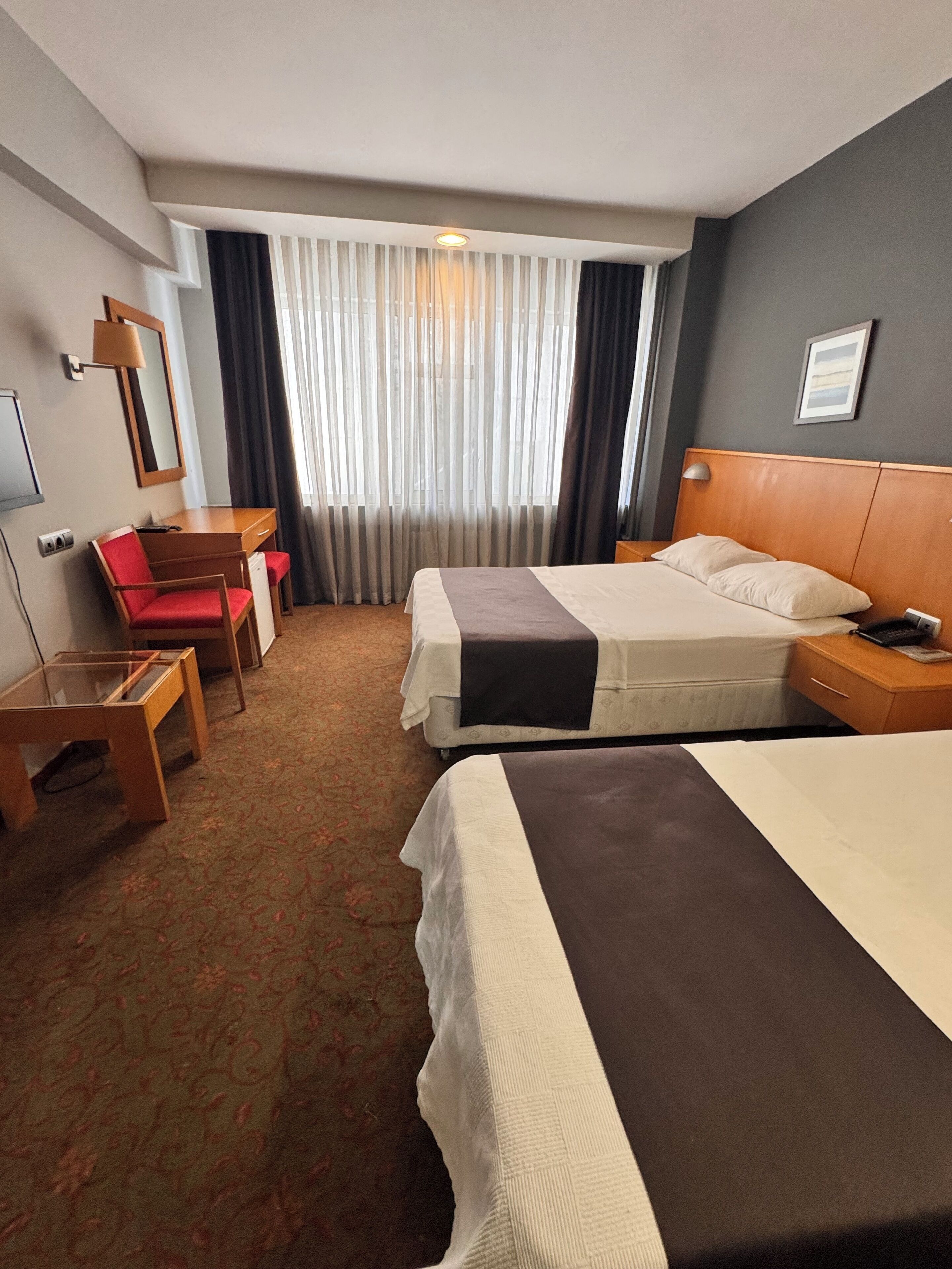 A hotel room with two beds, a desk, a chair, and a large window with curtains.