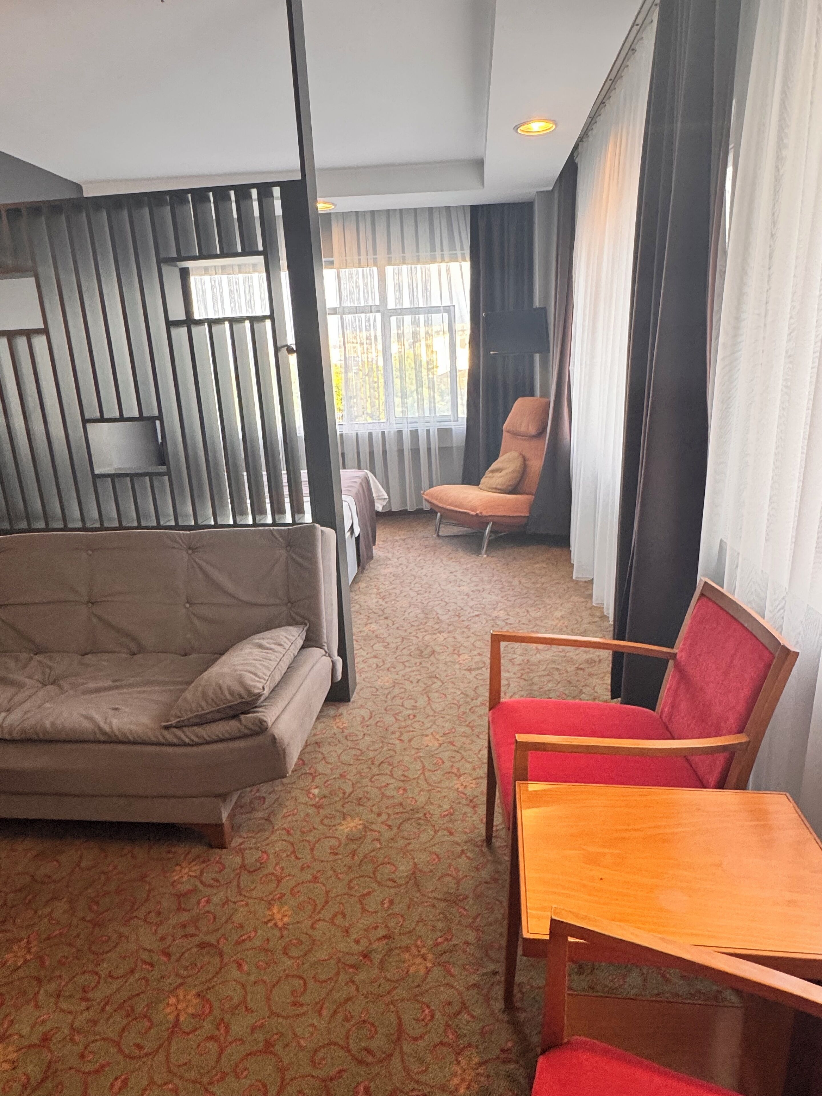 Una habitación de hotel con un sofá, una silla roja, una mesa de madera y una alfombra estampada.