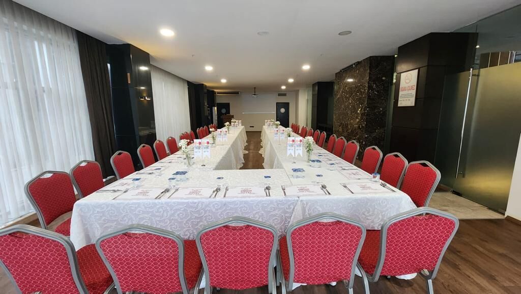 Un salón de conferencias con una larga mesa, sillas rojas y manteles blancos.