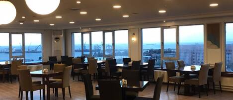 Uma ampla sala de jantar com várias mesas e cadeiras, grandes janelas com vista para a cidade e luminárias modernas.