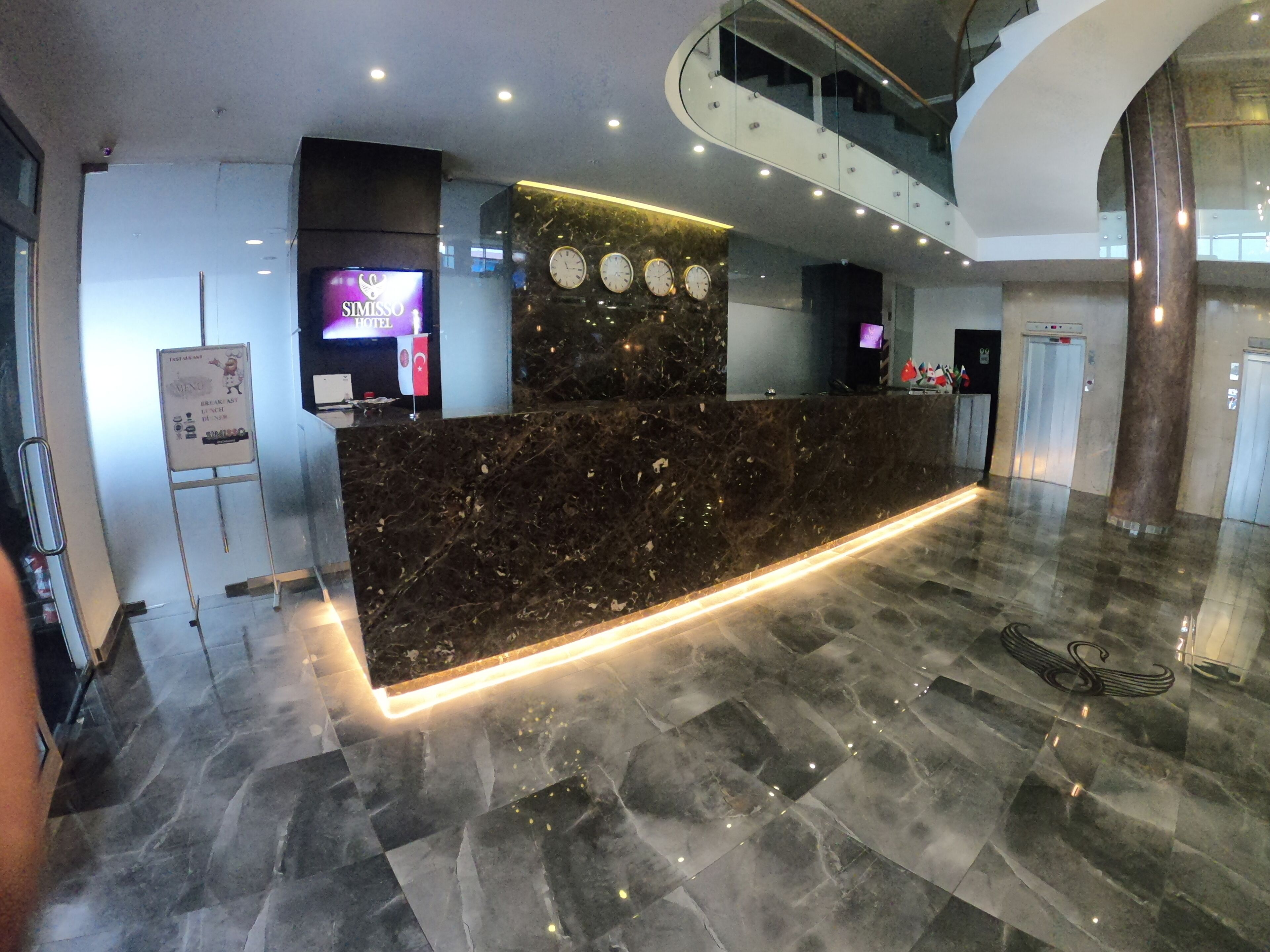 Una moderna zona de recepción de hotel con mostrador de mármol, relojes digitales y una escalera.