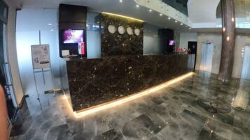 Una moderna zona de recepción de hotel con mostrador de mármol, relojes digitales y una escalera.
