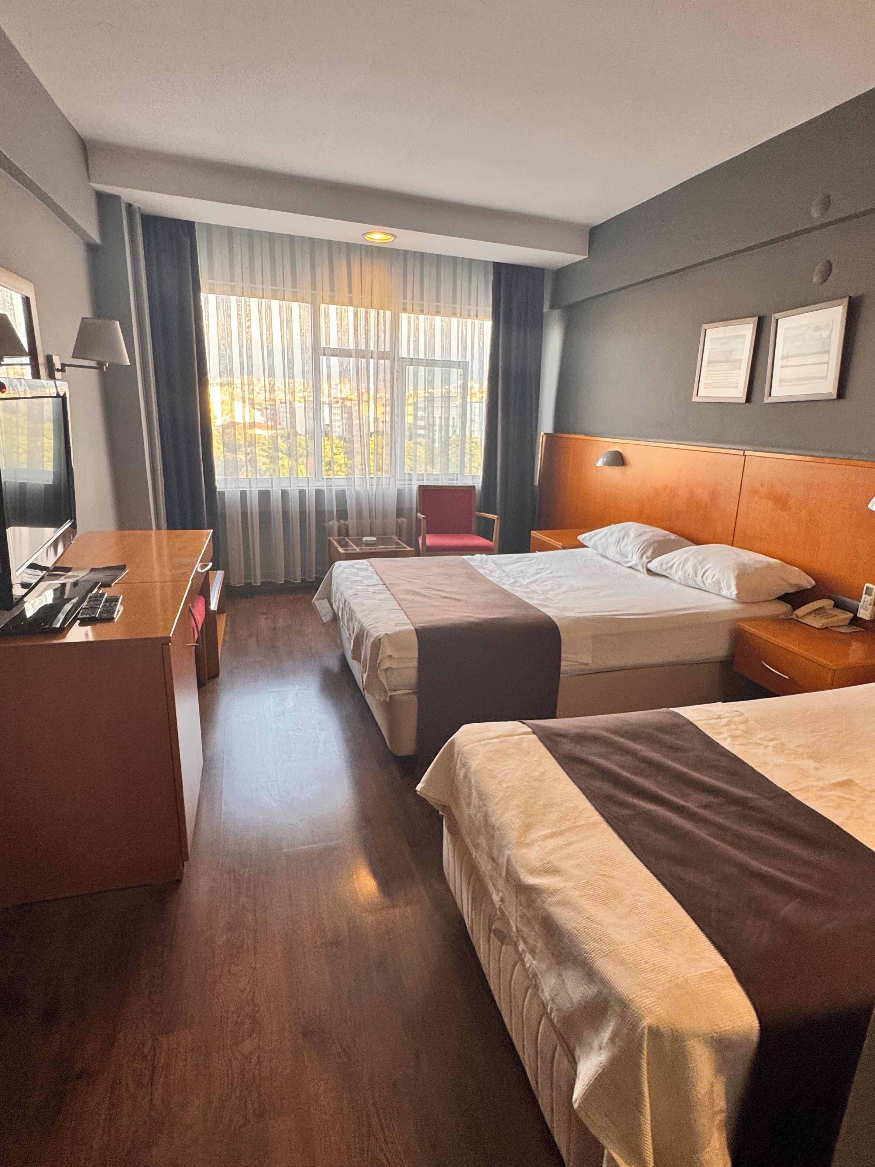 Habitación de hotel con dos camas, un escritorio, una silla, un televisor y un ventanal con cortinas.