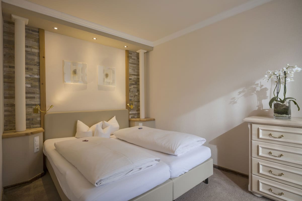 A neatly made double bed with white linens, a nightstand with a lamp, and a dresser with drawers.