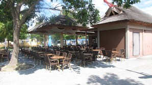 An outdoor dining area with tables and chairs, a thatched-roof structure, and a tree providing shade.