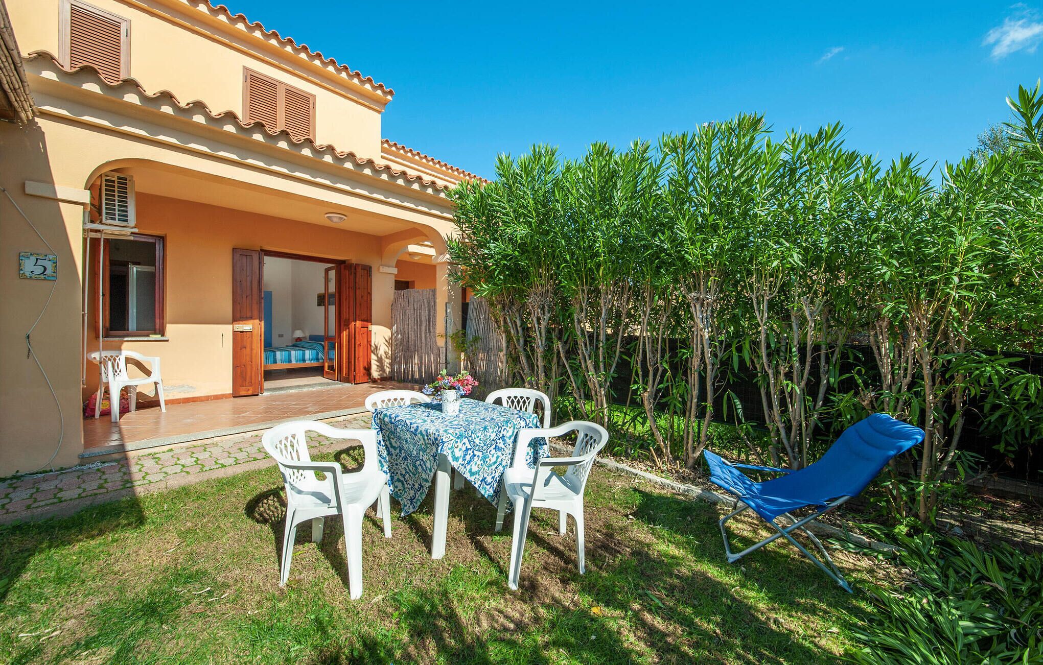 A two-story building with a patio area featuring a table and chairs, a blue lounge chair, and a garden with tall plants.