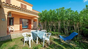 A two-story building with a patio area featuring a table and chairs, a blue lounge chair, and a garden with tall plants.