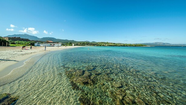 A beach with clear shallow waters, a few people, and a building in the background.