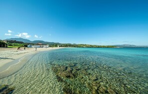 Une plage avec une eau claire et peu profonde, quelques personnes et un bâtiment en arrière-plan.