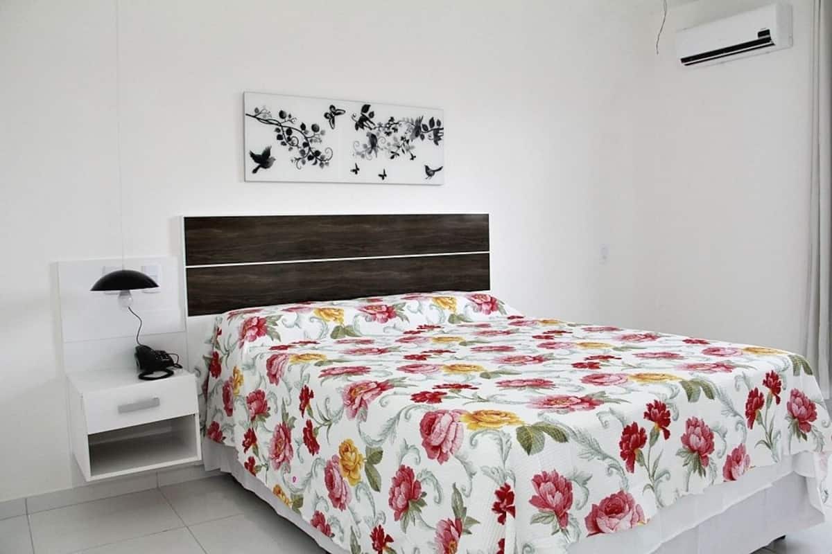 A bedroom with a bed, a bedside table with a lamp, a wall-mounted artwork, and a white air conditioning unit.