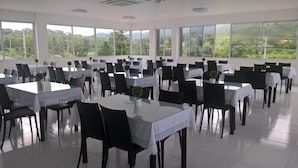 A large, well-lit dining area with multiple tables and chairs, large windows with a view of greenery, and a tiled floor. - France Hotel (Guarabira)