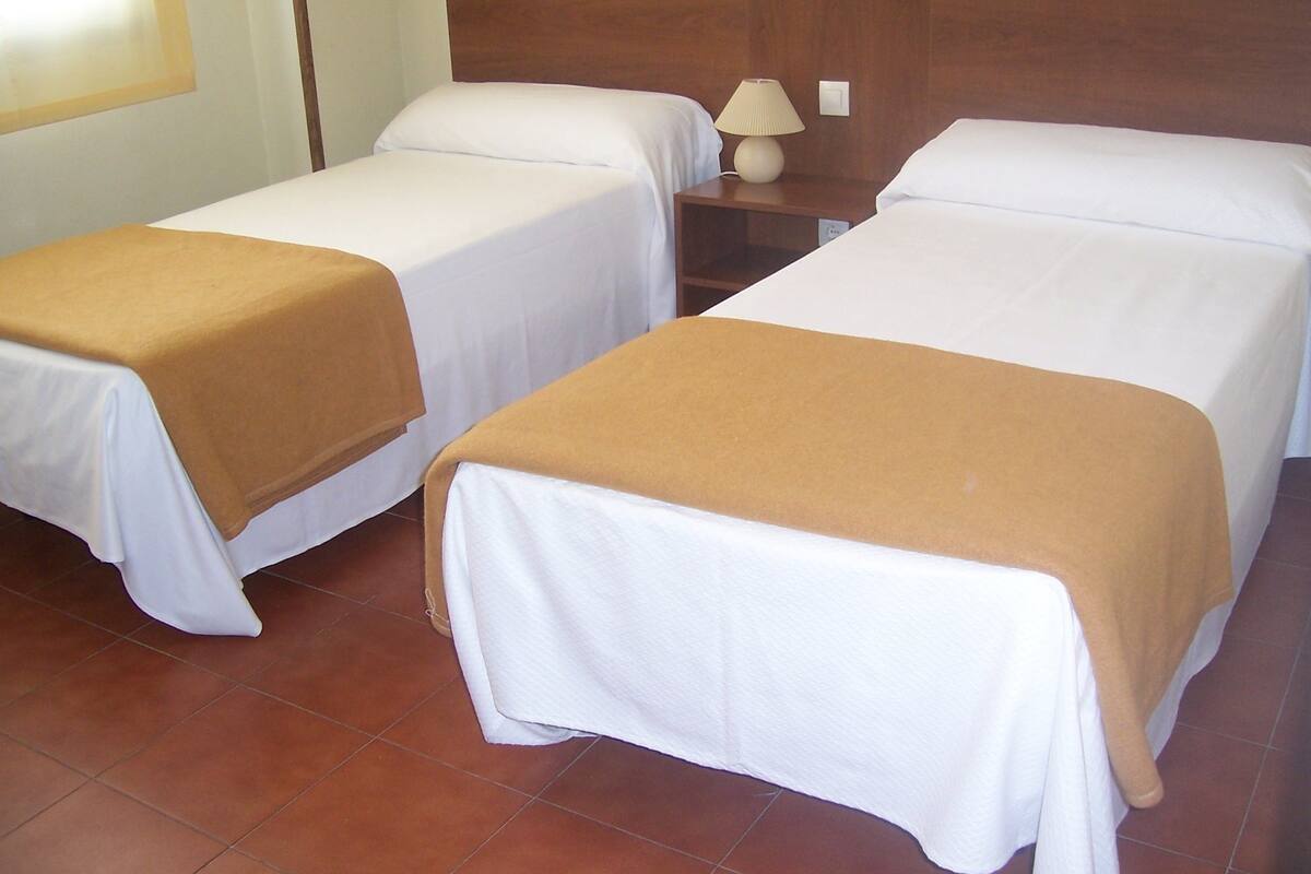 Dos camas individuales con sábanas blancas y mantas marrones, una mesita de noche con lámpara, y piso de baldosas.