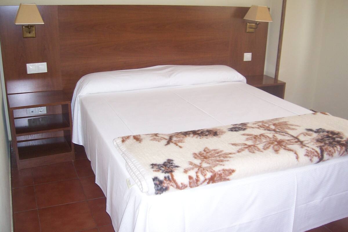 Una cama bien tendida con una manta floral, cabecera de madera, lámparas de noche y una mesita de noche de madera con toma de corriente.