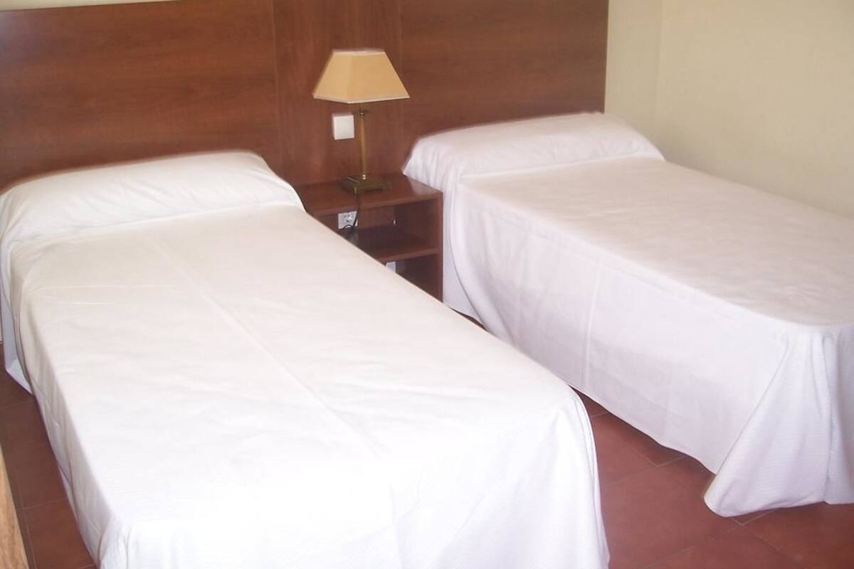 Una habitación con dos camas individuales, un cabecero de madera, una mesita de noche con lámpara y suelo de baldosas.