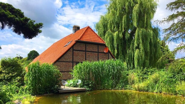 A traditional house with a red roof, surrounded by lush greenery and a pond. - Ferienhaus Villa EMG Düsseldorf Krefeld (Krefeld)