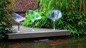 A wooden deck with three glass tables, surrounded by a pond with koi fish, and lush greenery. - Ferienhaus Villa EMG Düsseldorf Krefeld (Krefeld)