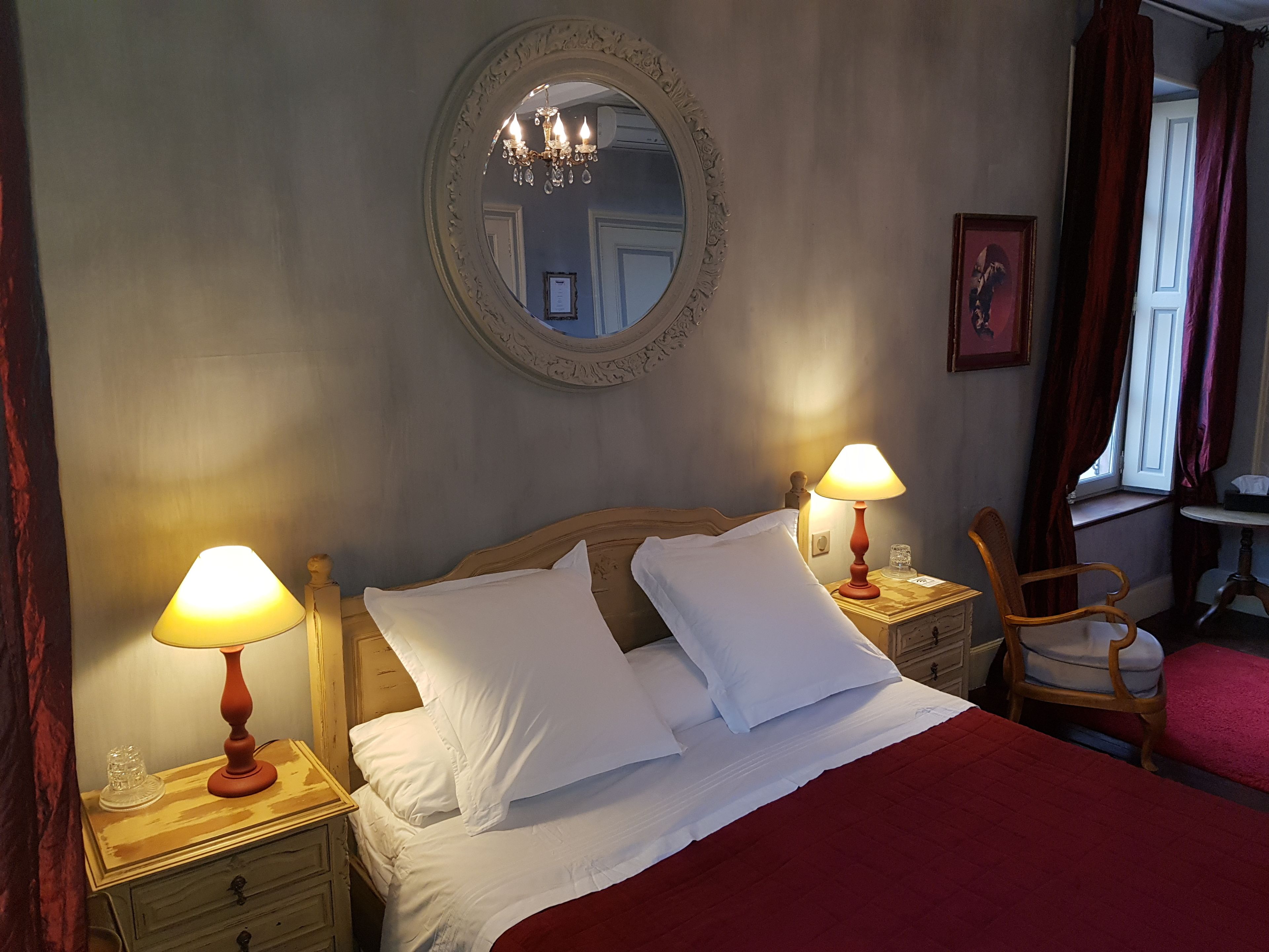 A neatly made bed with white linens, two bedside lamps, a wooden nightstand, a chair, and a mirror on the wall.