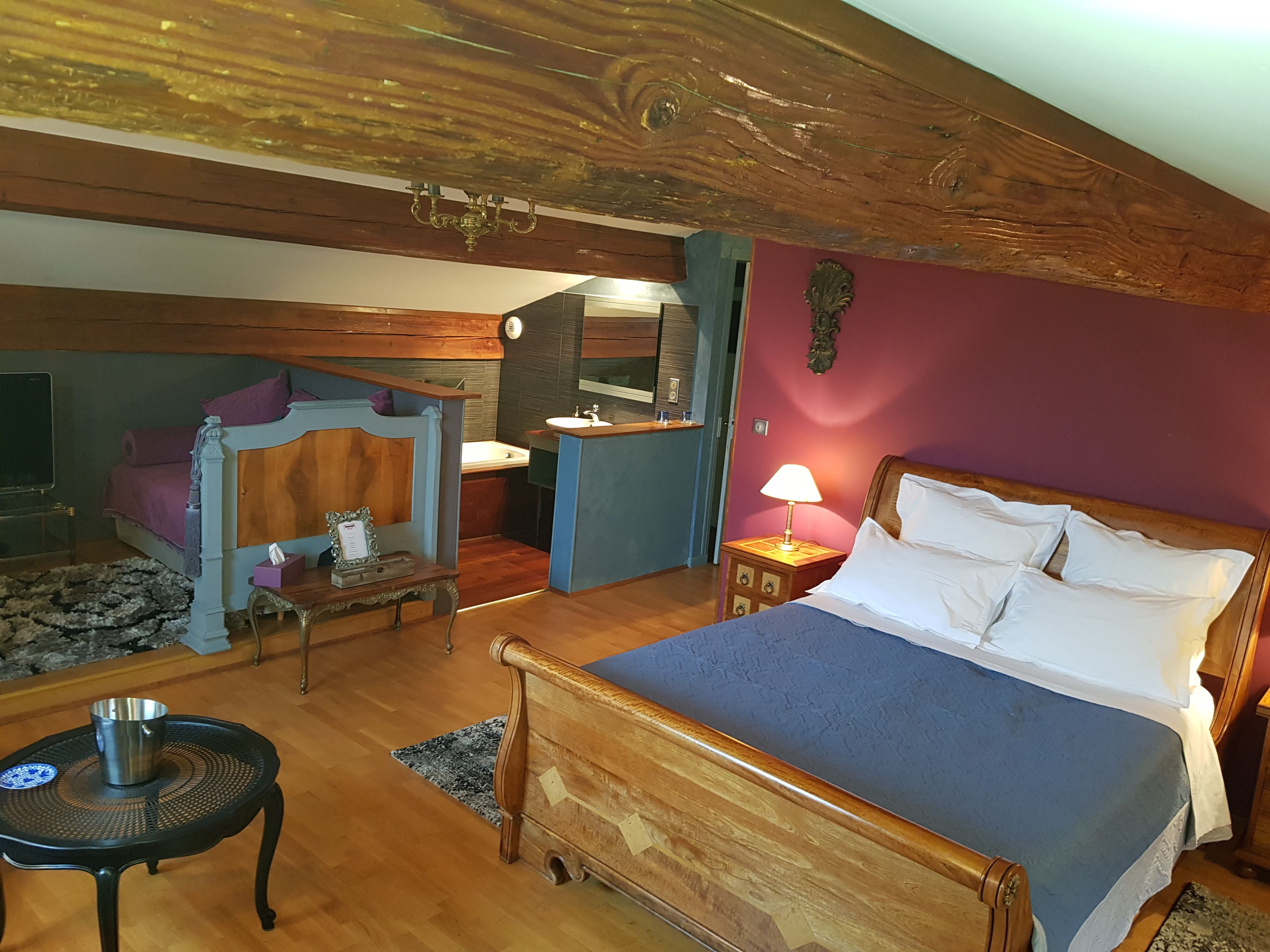 Un dormitorio con cama de madera, una mesita de noche con lámpara, una pequeña mesa con una taza, un banco y un baño con lavamanos y espejo.
