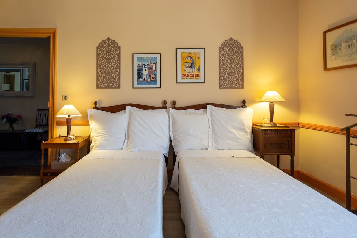 Una cama doble con sábanas blancas, dos mesitas de noche con lámparas, y tres cuadros en la pared.
