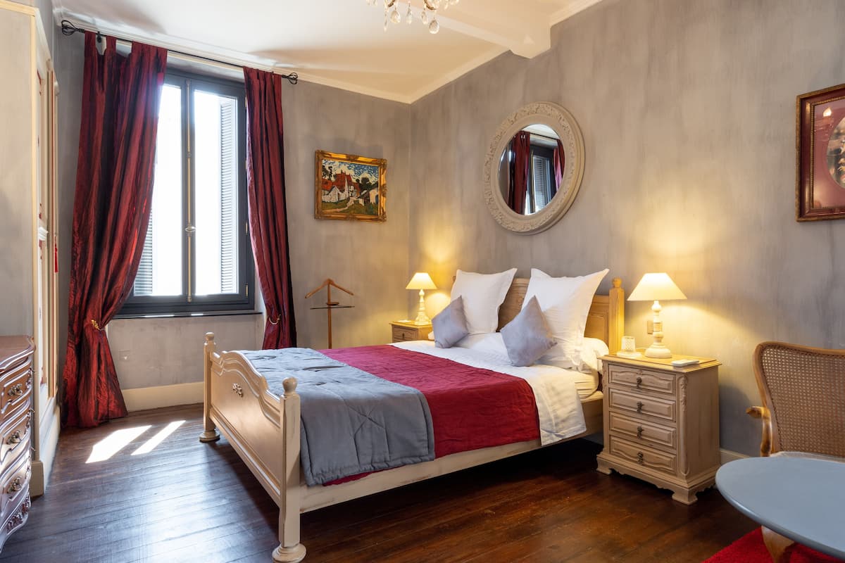 Un dormitorio con cama, mesitas de noche, una silla, un espejo y un cuadro en la pared.
