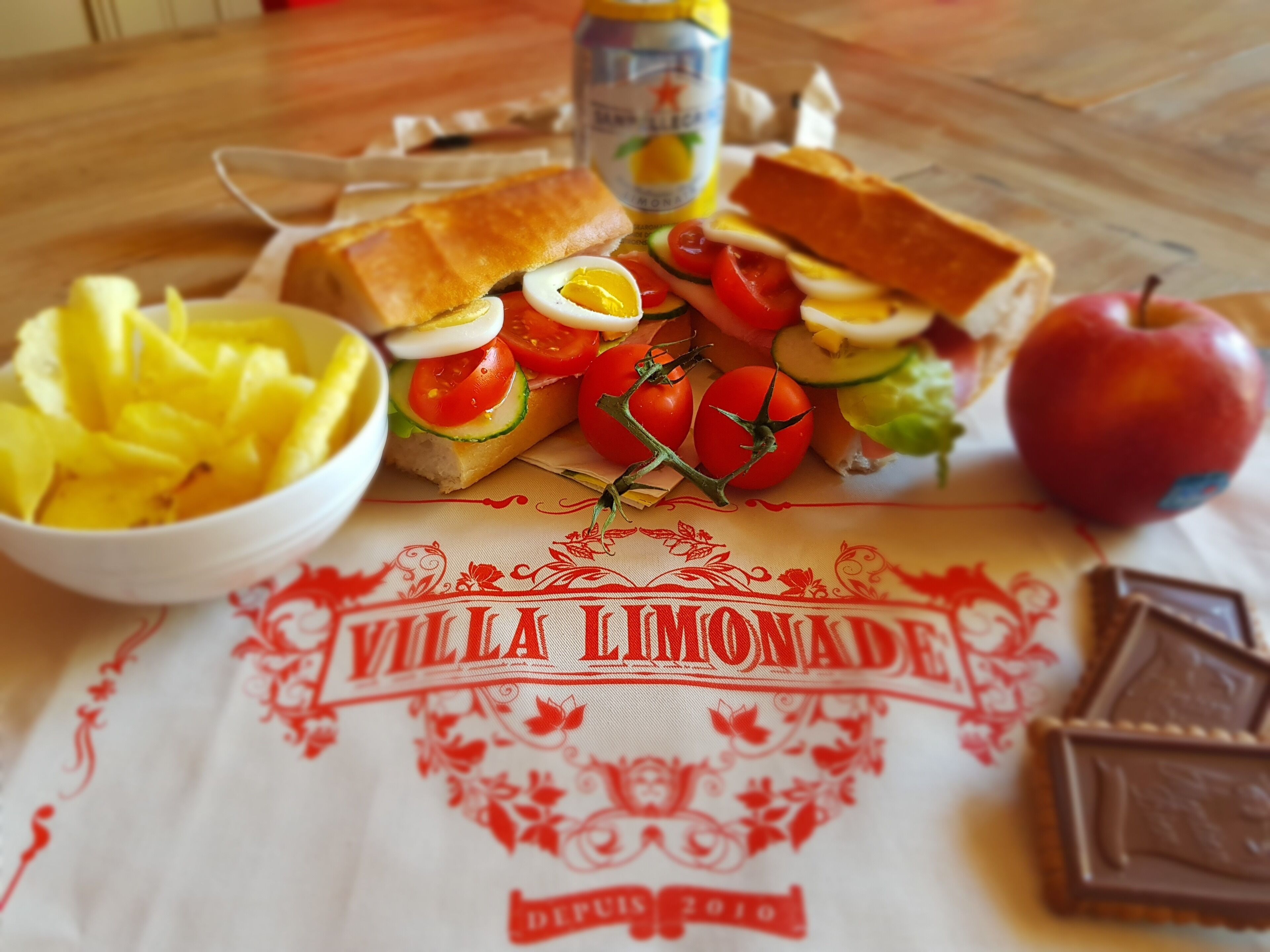 Un tavolo con un panino, pomodori, cetrioli, uova e una ciotola di ananas.