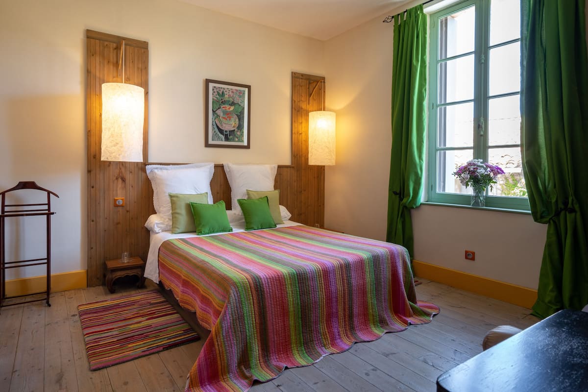 Un dormitorio con cabecera de madera, colcha a rayas de colores, dos lámparas fijadas a la pared, una mesita de noche de madera y una ventana con cortinas verdes.