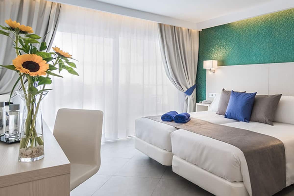A hotel room with a bed, bedside tables, a desk with a vase of flowers, and a view of the outdoors.