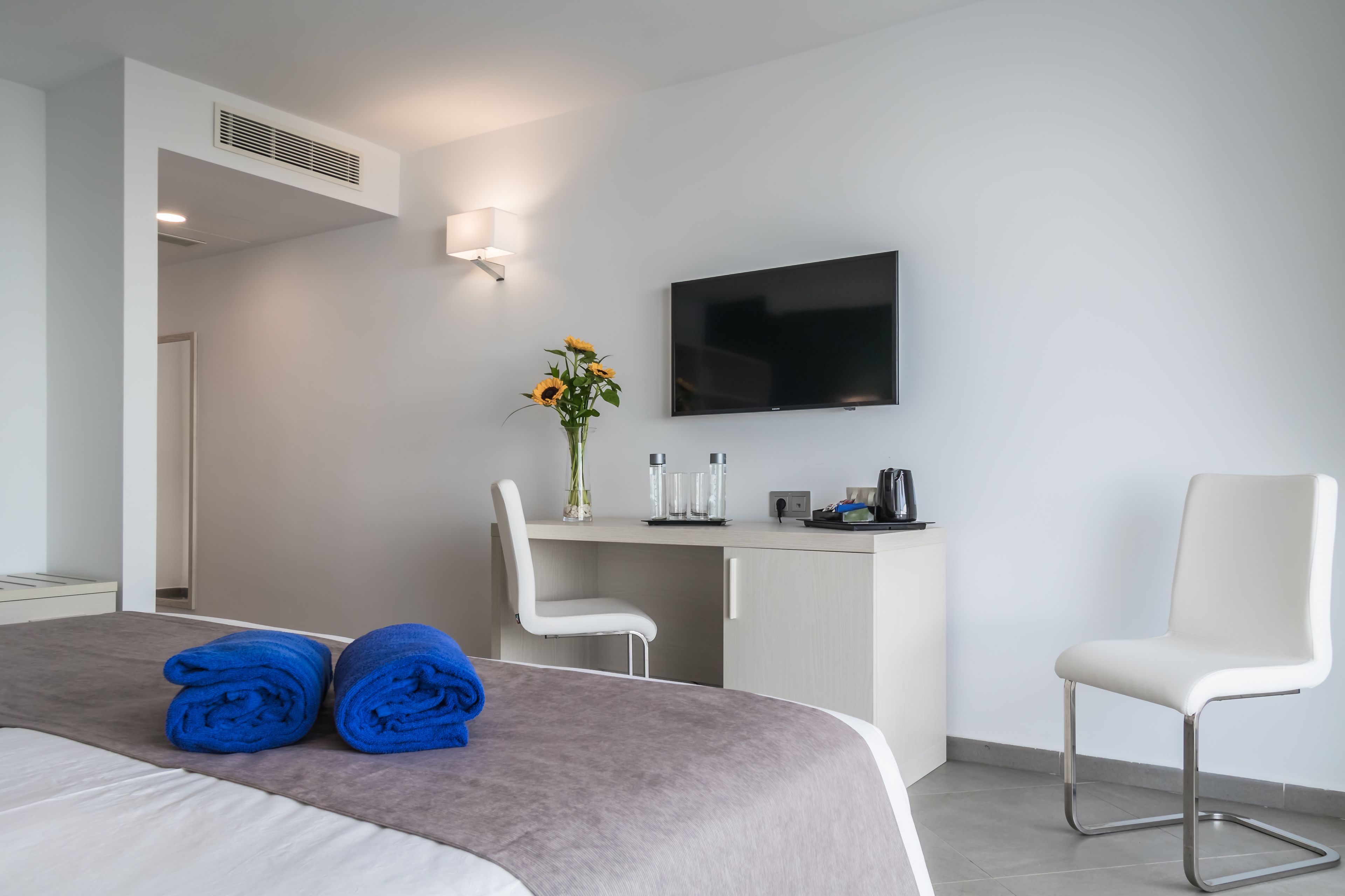 A modern hotel room with a bed, two white chairs, a desk with a flat-screen TV, a vase with flowers, and a coffee machine.
