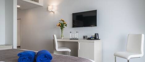 A modern hotel room with a bed, two white chairs, a desk with a flat-screen TV, a vase with flowers, and a coffee machine.