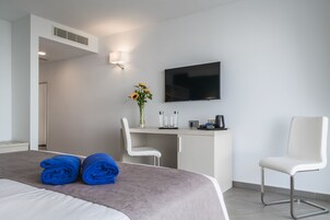 A modern hotel room with a bed, two white chairs, a desk with a flat-screen TV, a vase with flowers, and a coffee machine.