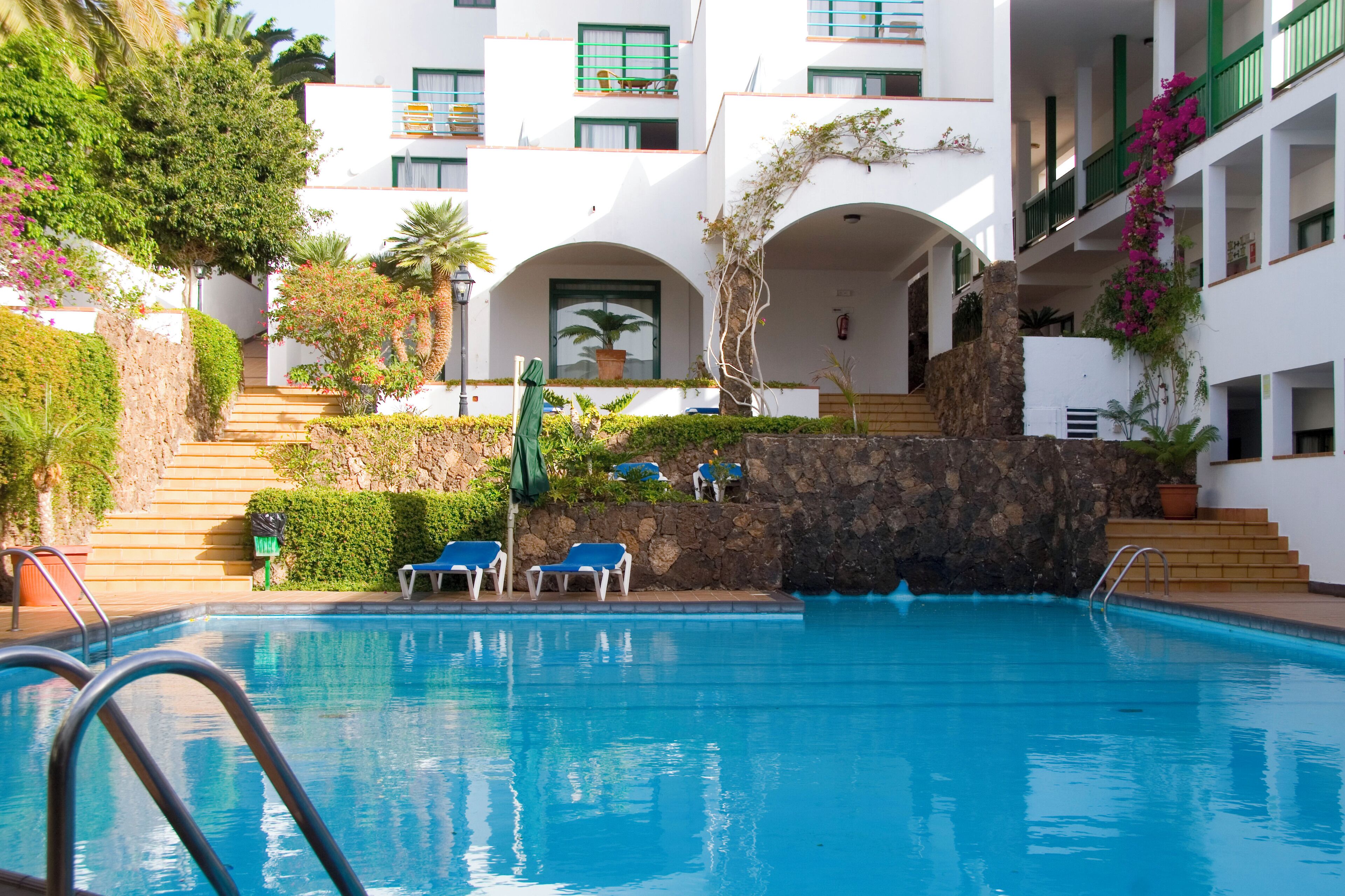A swimming pool with blue water, surrounded by white buildings with green balconies and arches.