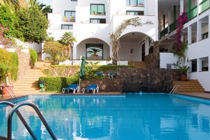 Una piscina con agua azul, rodeada de edificios blancos con balcones y arcos verdes.