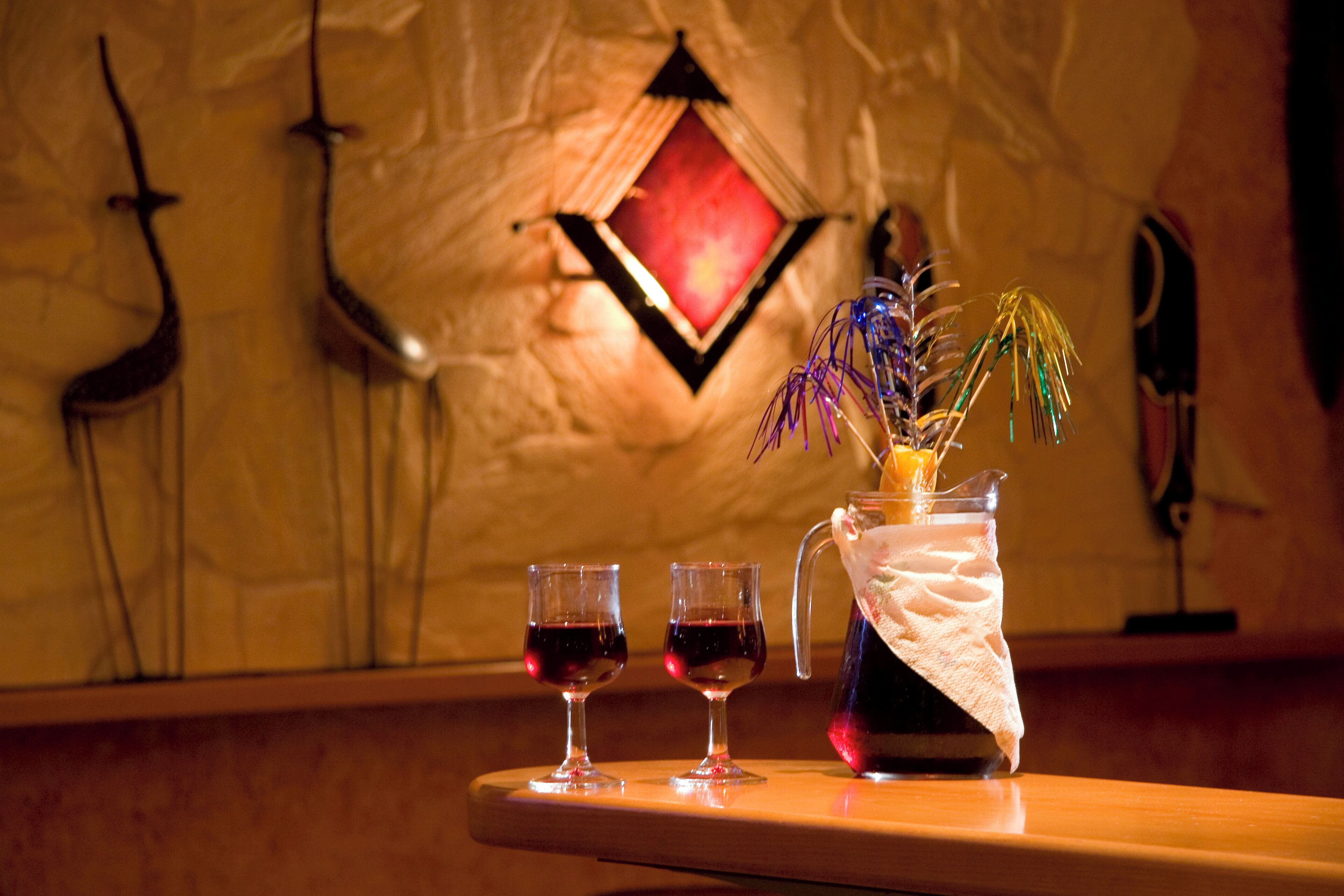 A bar area with two glasses of red wine, a pitcher with a decorative plant, and a wall with a diamond-shaped mirror and wall-mounted decorations.
