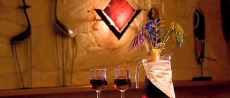 A bar area with two glasses of red wine, a pitcher with a decorative plant, and a wall with a diamond-shaped mirror and wall-mounted decorations.