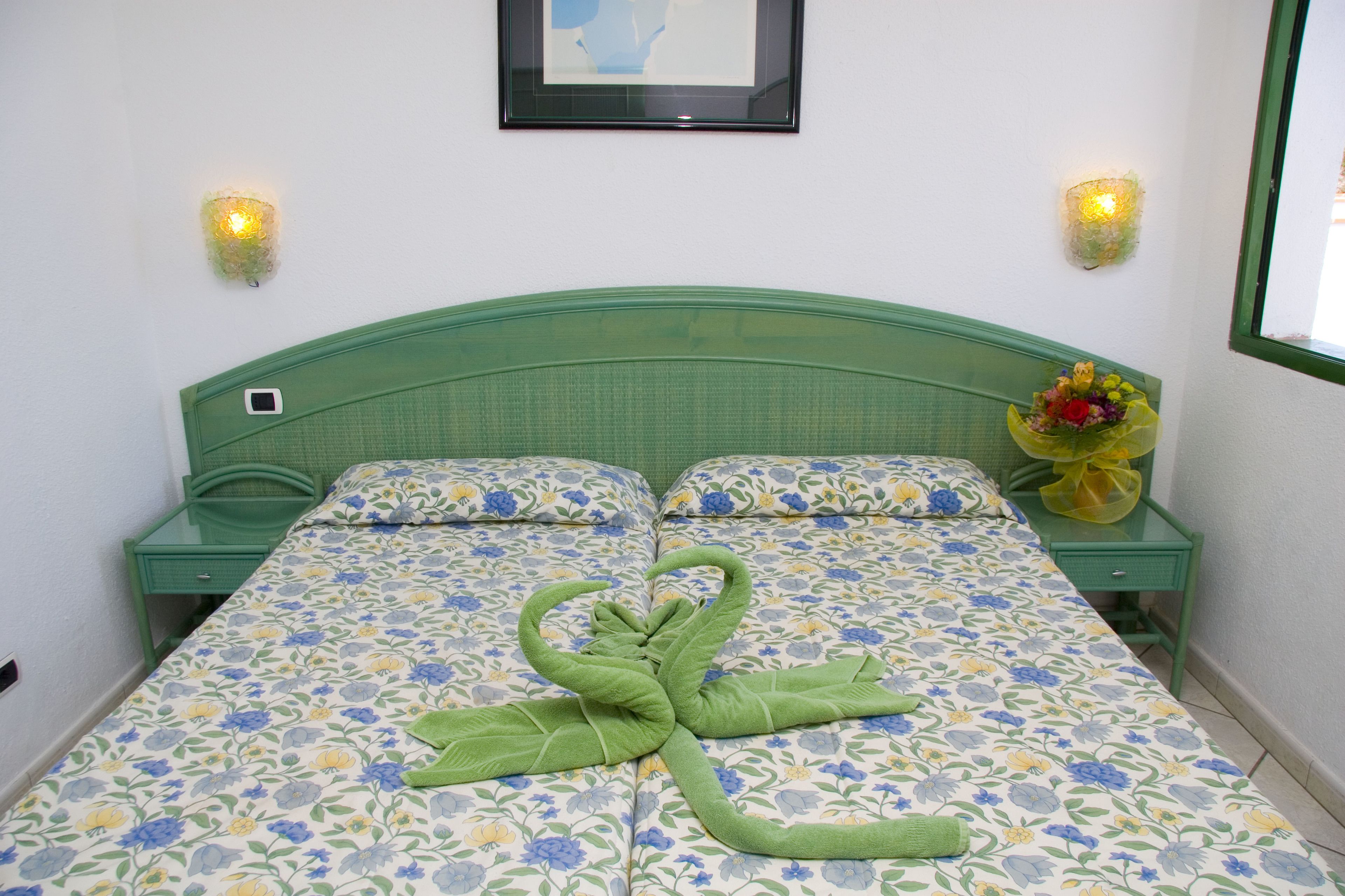 Una cama con una colcha floral, un cabecero verde y un ramo de flores sobre una mesita de noche.