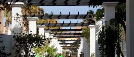 A covered walkway with white columns and a pergola overhead, surrounded by lush greenery and potted plants.