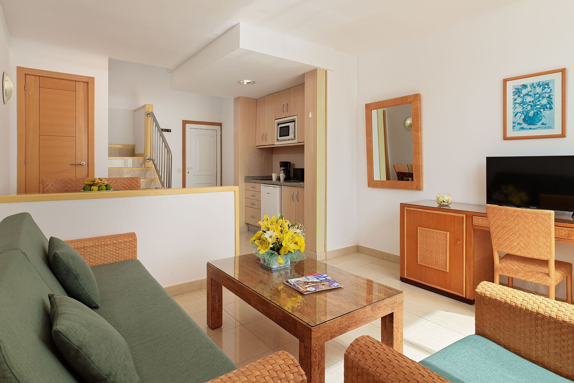 A living room with a sofa, a coffee table with flowers, a television, and a kitchen area with cabinets and appliances.