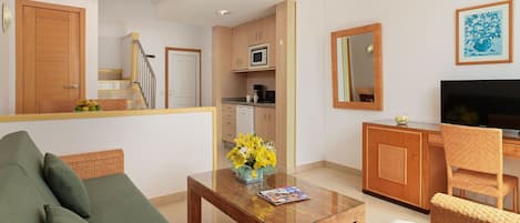 A living room with a sofa, a coffee table with flowers, a television, and a kitchen area with cabinets and appliances.