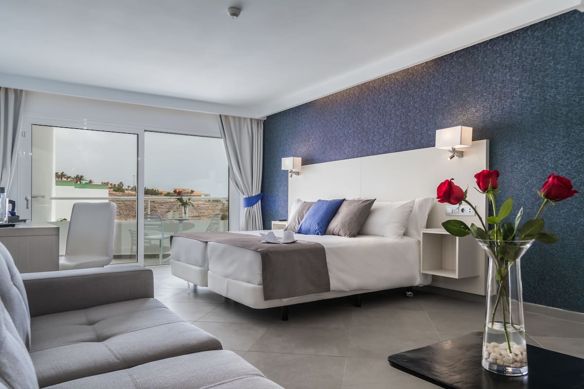 A modern hotel room with a bed, a sofa, a glass door leading to a balcony, and a vase with red roses.