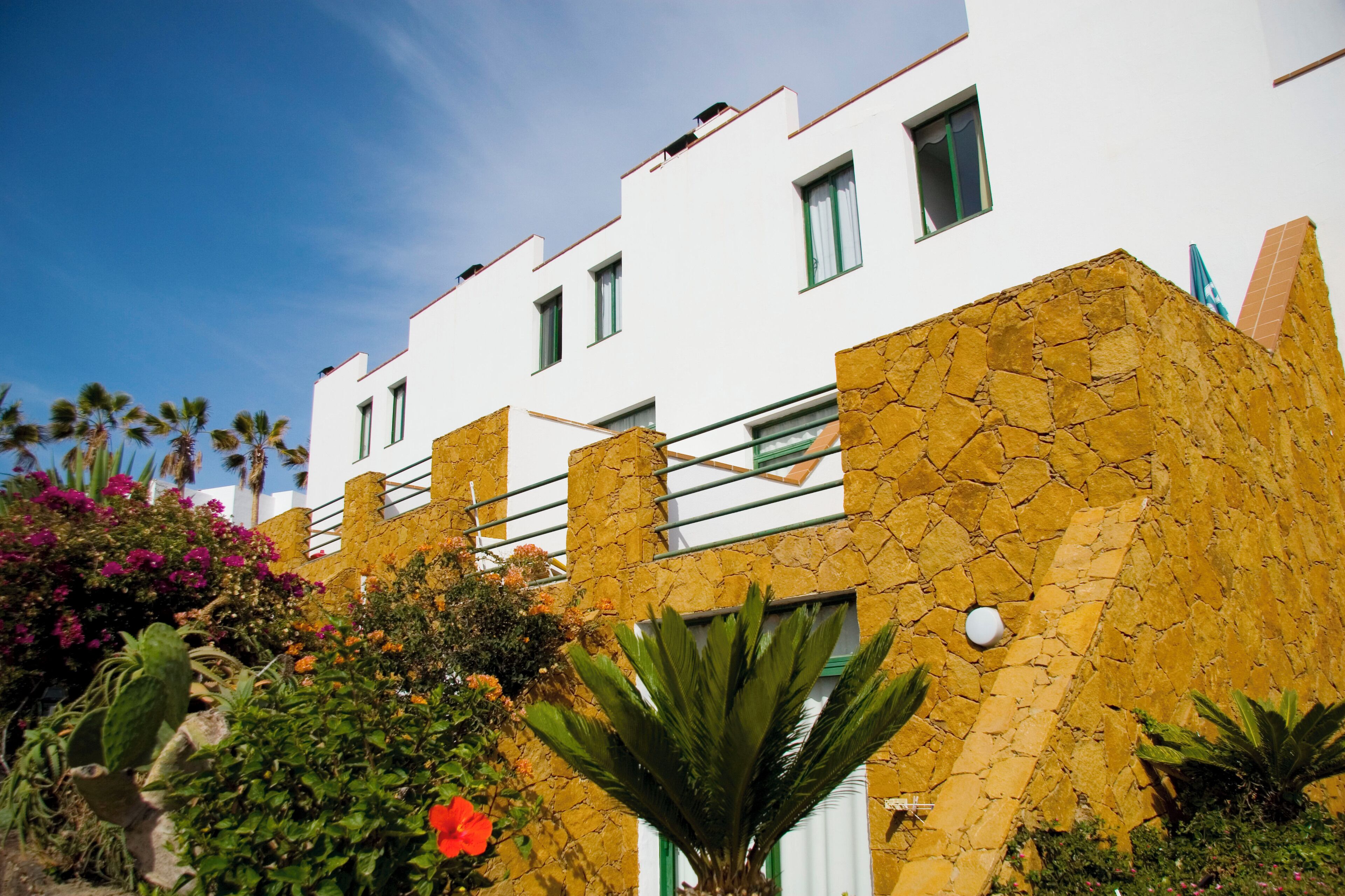 Un bâtiment moderne aux murs blancs et à la façade en pierre, agrémenté de balcons et surplombant un ciel d’un bleu éclatant.