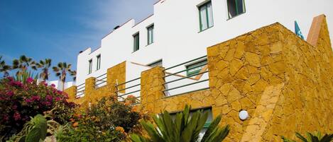 A modern building with white walls and a stone facade, featuring balconies and a clear blue sky.