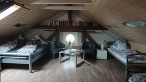 A cozy attic bedroom with a loft bed, a small table, and a chair. There is a window with a view of the sky, and books are visible on the shelves.