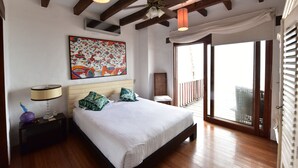 A bedroom with a large bed, wooden beams, a ceiling fan, a painting, a nightstand, and a view through a sliding door. - Casa Baronesa (Puerto Villamil)